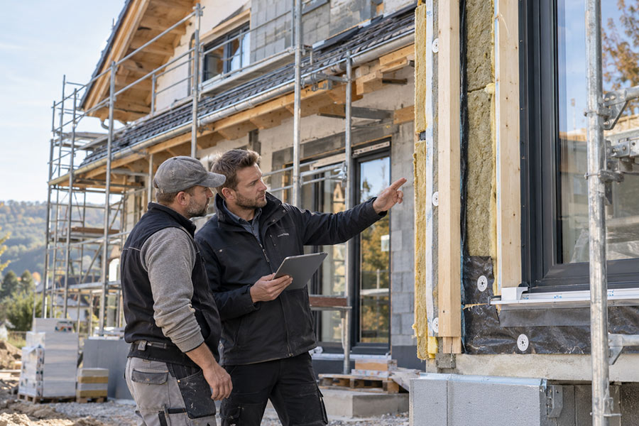 Zwei Fachleute auf der Baustelle begutachten die Dämmung einer Fassade, einer zeigt auf das freiliegende Dämmmaterial